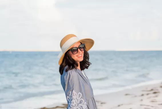 Side view of a barefoot woman in a flowing blue embroidered beach dress and straw hat walking along the sand under a cloudy sky with text crediting her outfit.